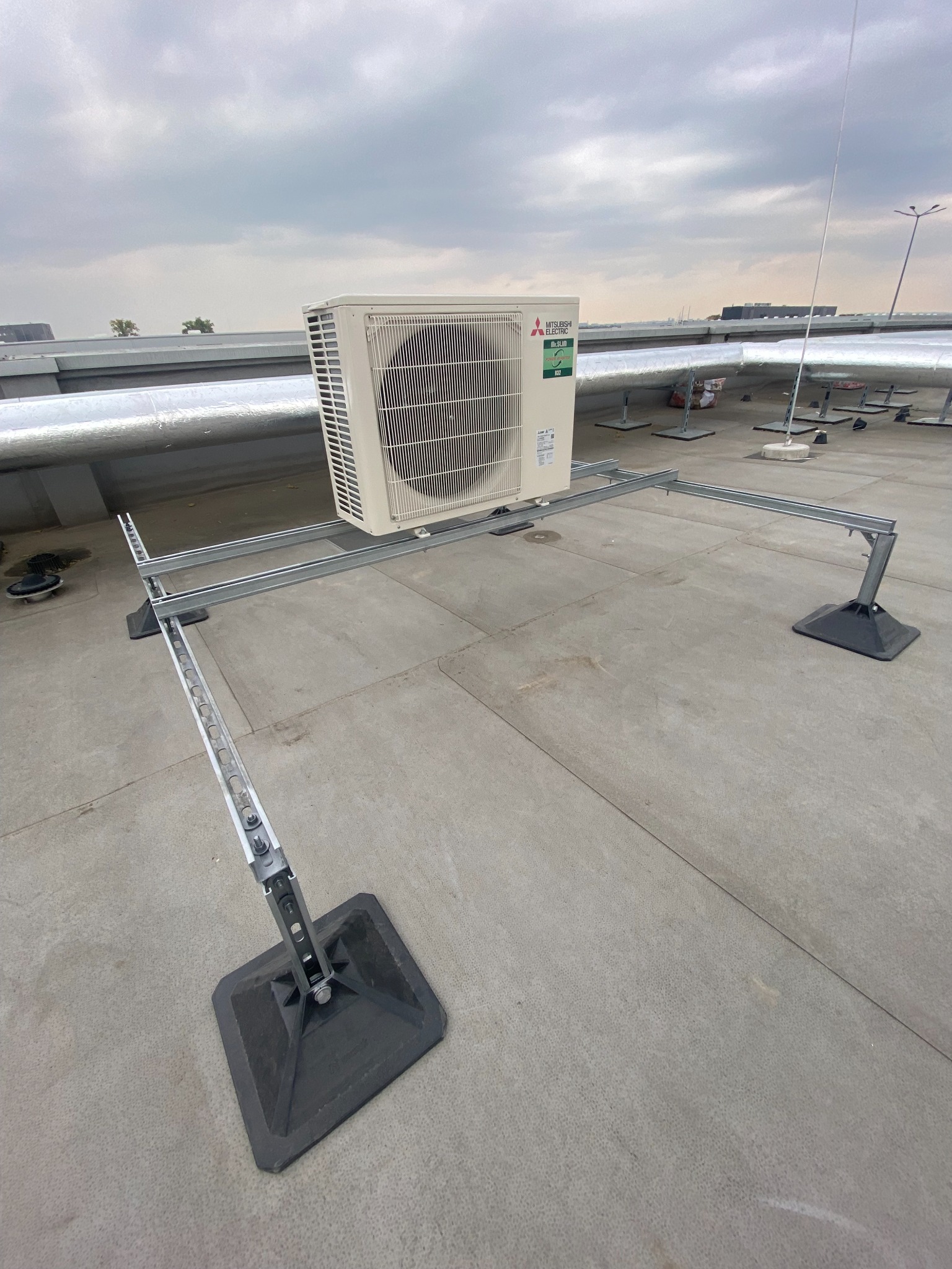 Klimatyzator Mitsubishi Electric zamontowany na metalowej konstrukcji wsporczej na płaskim dachu budynku przemysłowego, widoczne rury wentylacyjne w tle i pochmurne niebo.