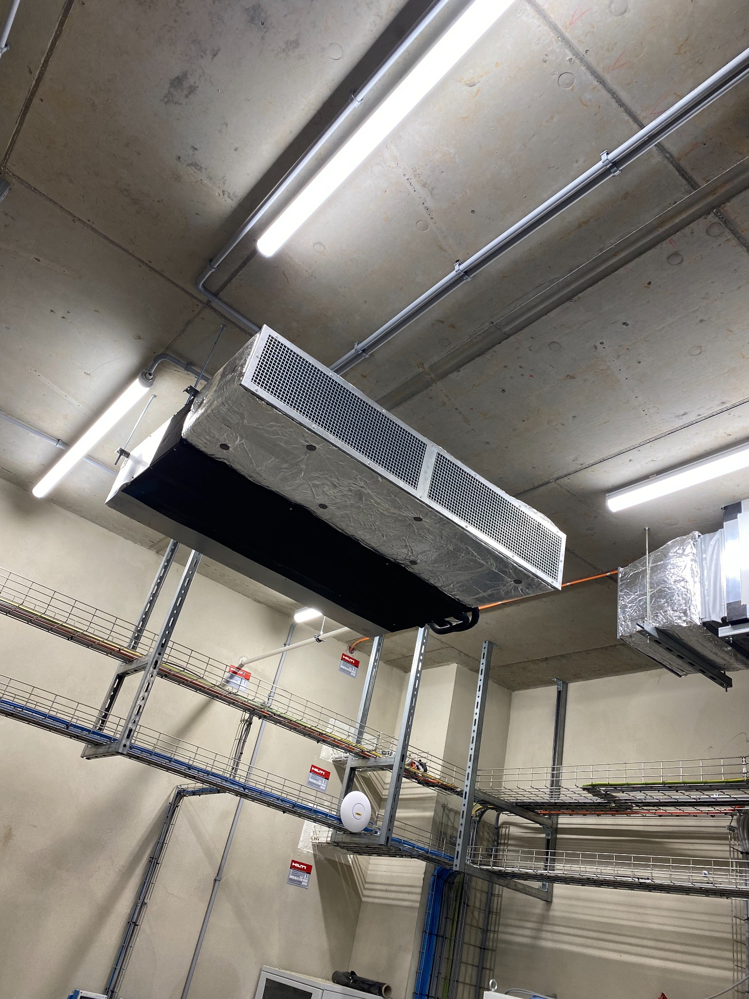 Zawieszony pod sufitem system wentylacyjny z metalową kratką i folią izolacyjną, widoczne korytka kablowe i oświetlenie przemysłowe