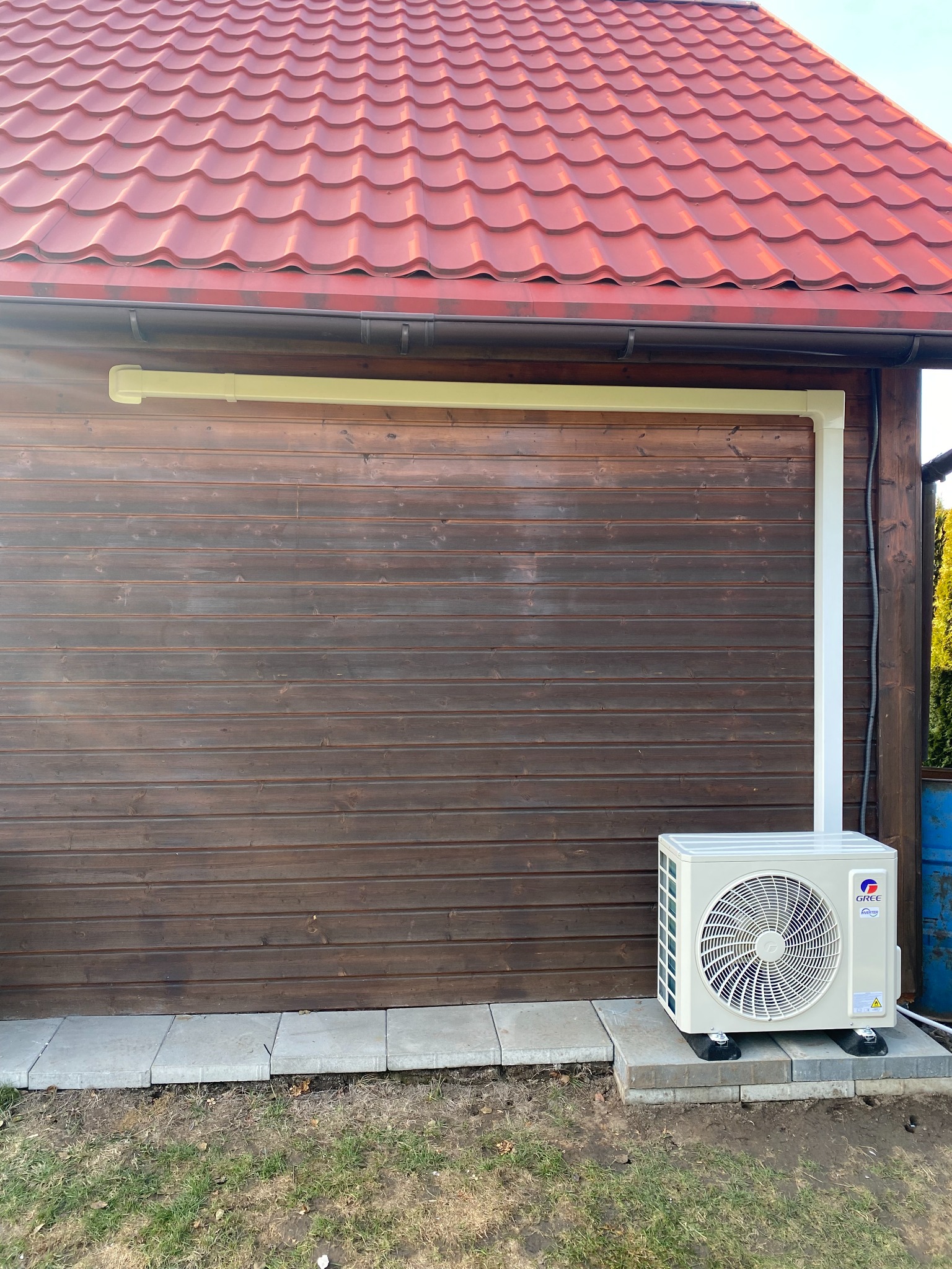 Zewnętrzna jednostka klimatyzacji marki Gree zamontowana na szarych płytach chodnikowych przy drewnianej ścianie budynku z czerwonym dachem.