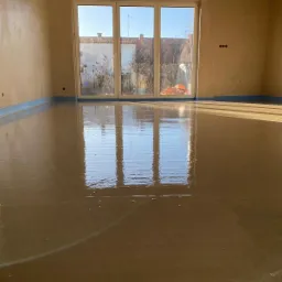 Świeżo wylana, gładka i błyszcząca posadzka w jasnym pomieszczeniu z dużym oknem, zabezpieczona niebieską taśmą przy ścianach.