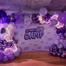 Scena udekorowana fioletowo-białymi balonami z napisem 'So Sweet Camp' na tle, oświetlona fioletowym światłem.
