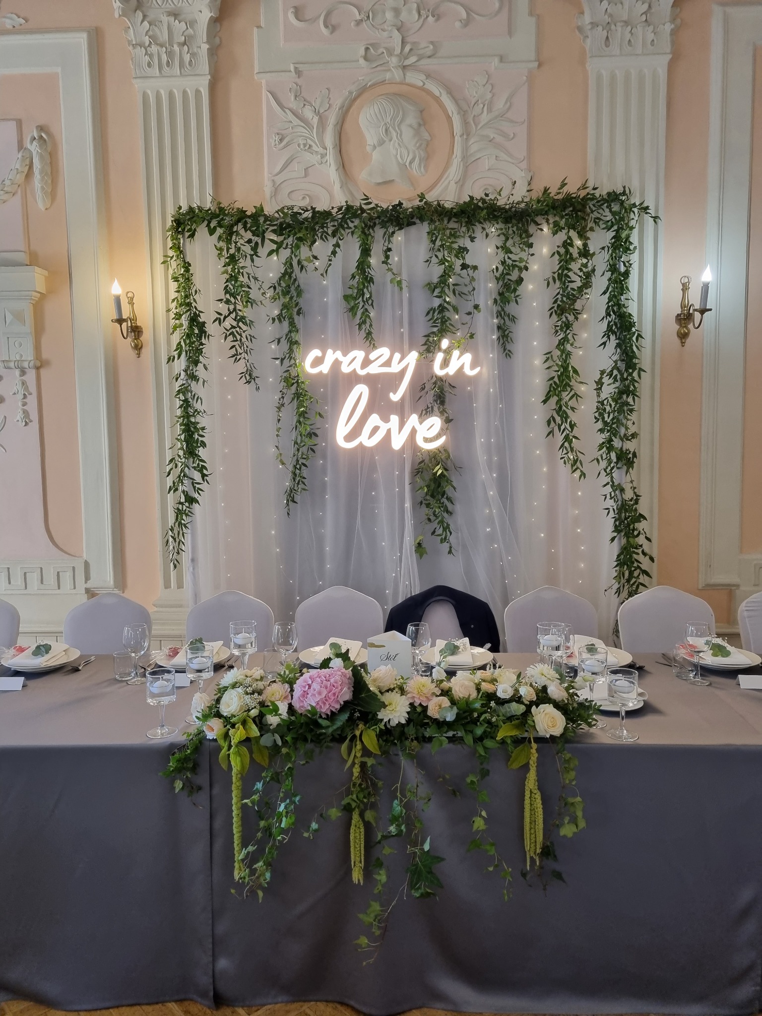 Uroczysty stół weselny z szarą tkaniną, kwiatową kompozycją z różowych hortensji i białych róż, nakryciami dla gości oraz neonowym napisem 'crazy in love' na tle białej, prześwitującej zasłony...