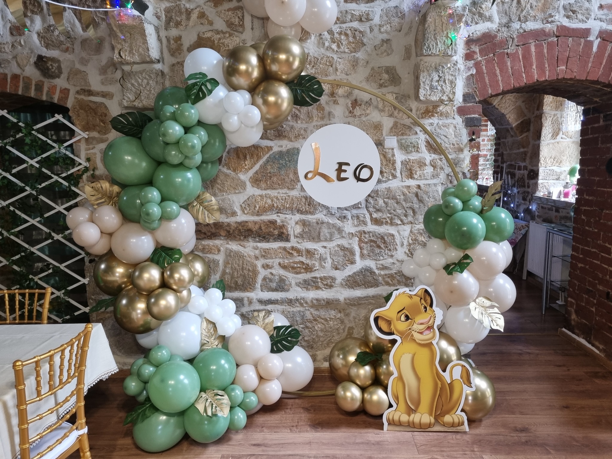 Dekoracja balonowa w odcieniach zieleni, bieli i złota z motywem Lwa, ustawiona na tle kamiennej ściany i łuku z cegły.