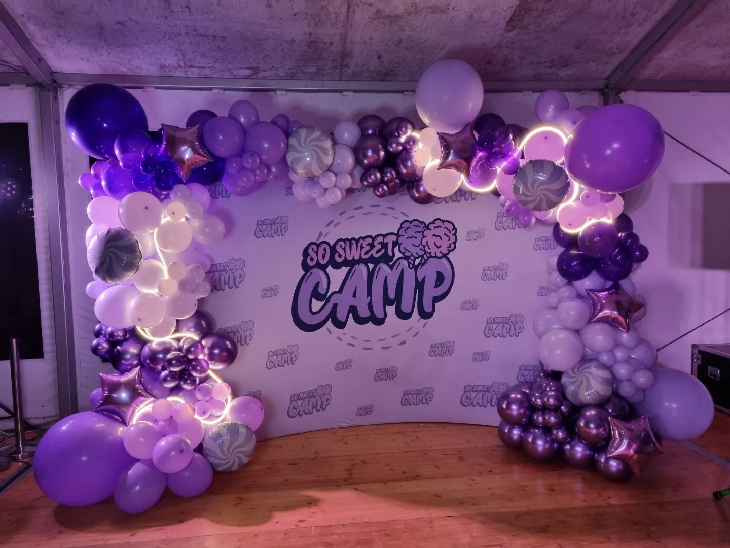 Scena udekorowana fioletowo-białymi balonami z napisem 'So Sweet Camp' na tle, oświetlona fioletowym światłem.