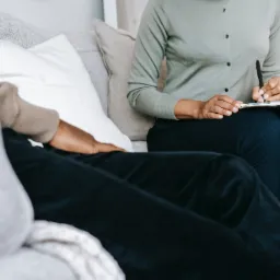 Fizjoterapeutka w zielonym swetrze siedzi na kanapie obok pacjenta i notuje informacje na clipboardzie.