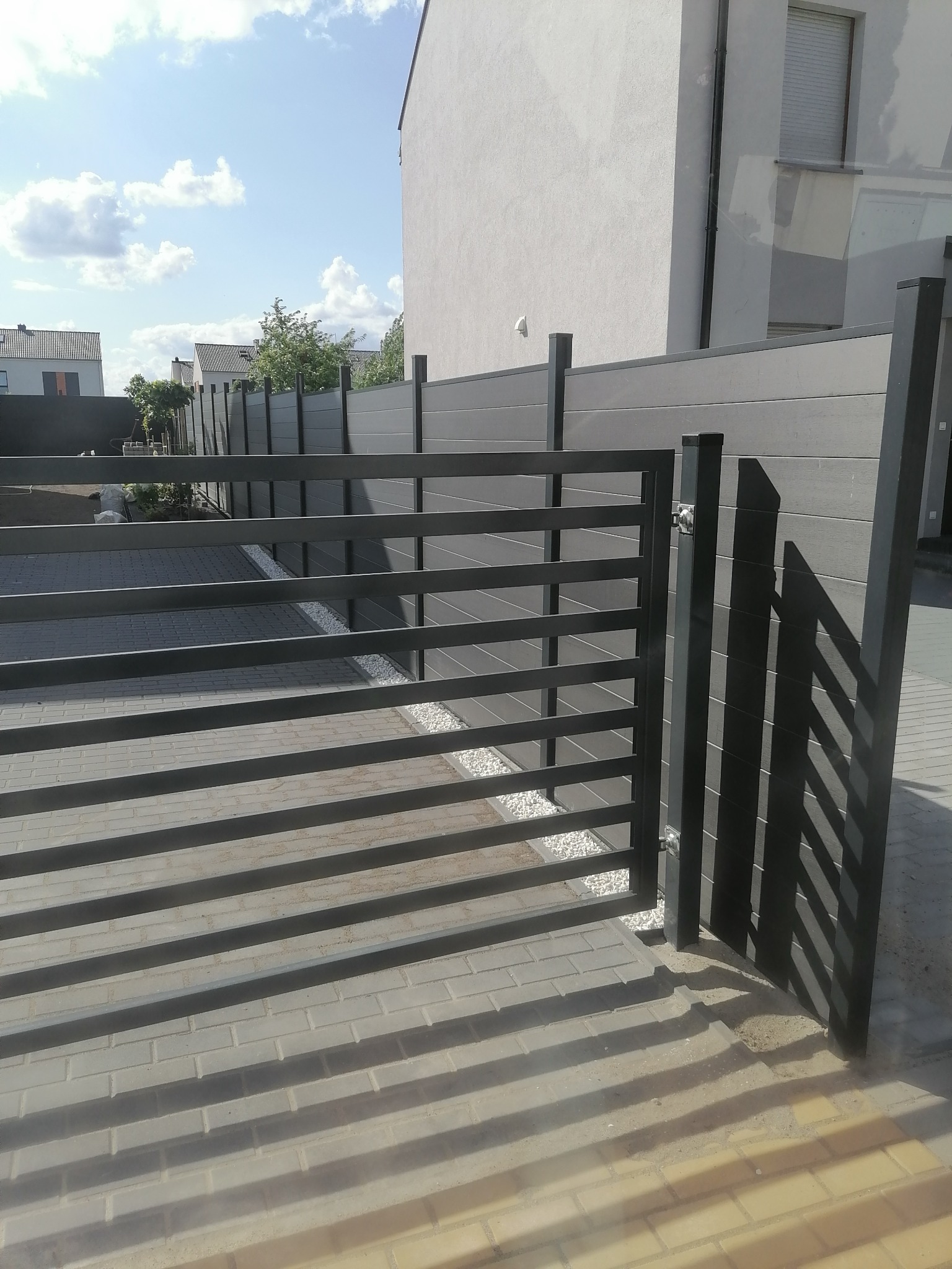 Nowoczesna, grafitowa brama furtkowa z poziomych profili, zamykająca wejście do posesji z szarym ogrodzeniem panelowym i kostką brukową w tle.