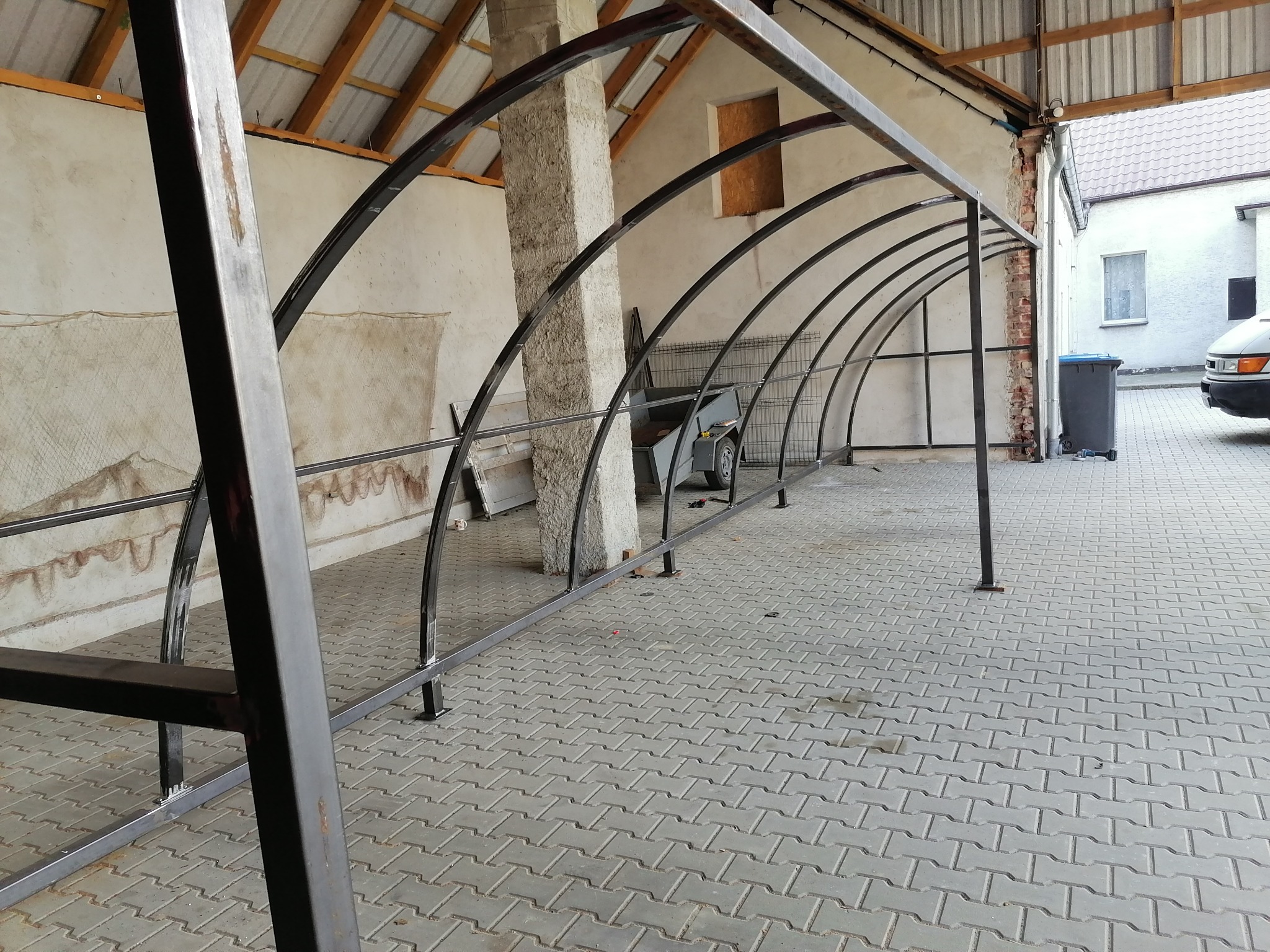 Konstrukcja z metalowych profili tworząca zadaszenie, widoczna na tle budynku gospodarczego i kostki brukowej.
