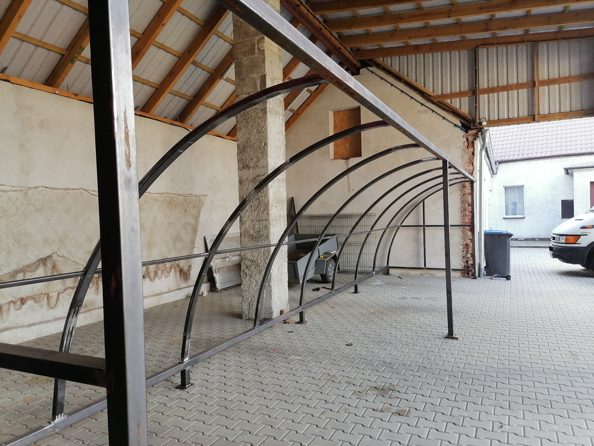 Konstrukcja z metalowych łuków w trakcie budowy, widoczna przyczepka w tle, surowy stan wykończenia.