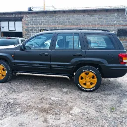 Czarny Jeep Grand Cherokee z matowym lakierem i pomarańczowymi felgami na tle szarego budynku warsztatowego z otwartą bramą.