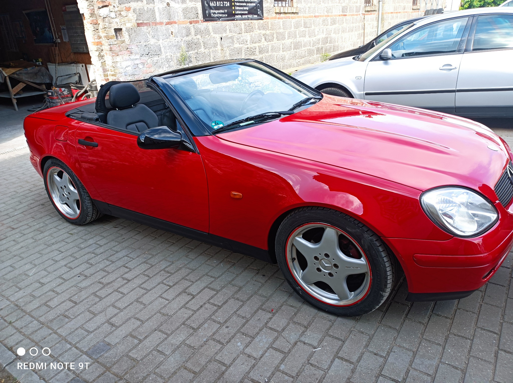 Czerwony kabriolet Mercedes SLK z czarnym dachem i srebrnymi felgami AMG, zaparkowany na brukowanym podjeździe przed szarą ceglaną ścianą warsztatu z widocznym szyldem reklamowym.