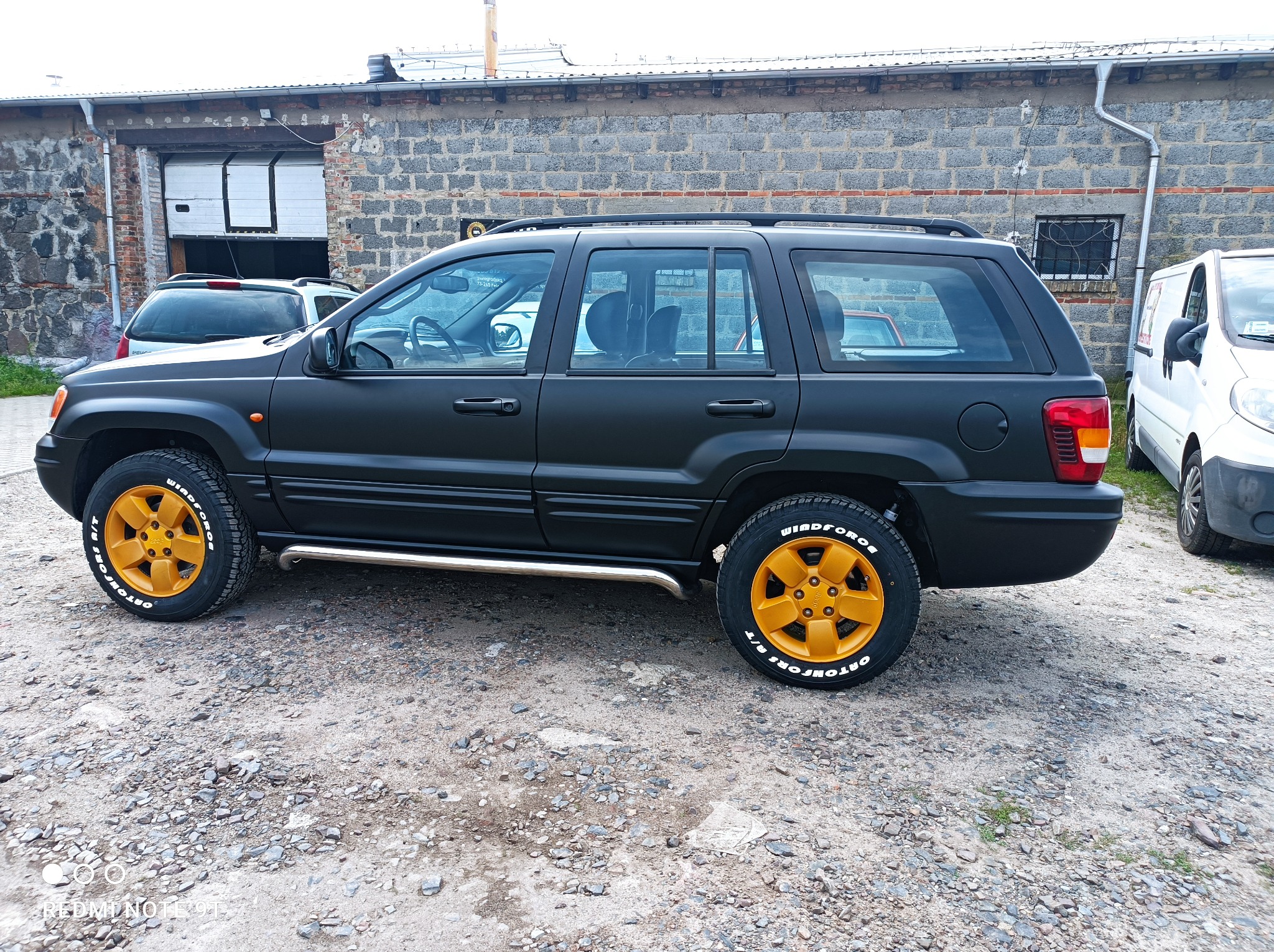 Czarny Jeep Grand Cherokee z matowym lakierem i pomarańczowymi felgami na tle szarego budynku warsztatowego z otwartą bramą.