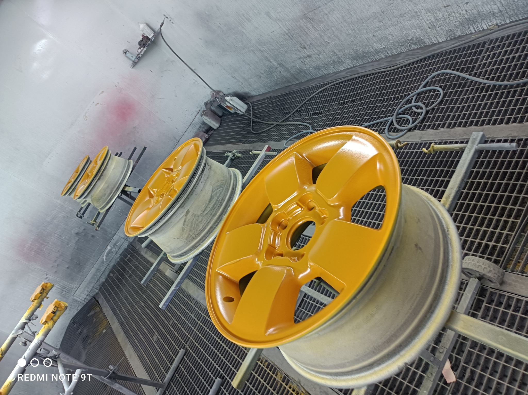 Żółte felgi samochodowe po lakierowaniu, ustawione w lakierni na metalowym ruszcie, widoczne narzędzia i przewody lakiernicze.