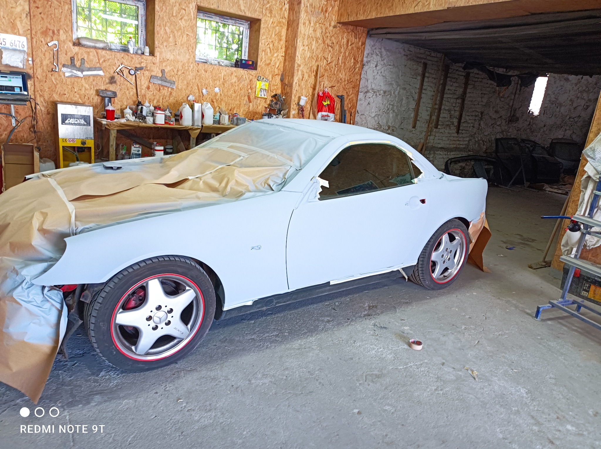 Samochód Mercedes SLK w trakcie lakierowania, częściowo zakryty papierem maskującym, widoczne felgi z czerwonym akcentem, warsztat z narzędziami i akcesoriami w tle.