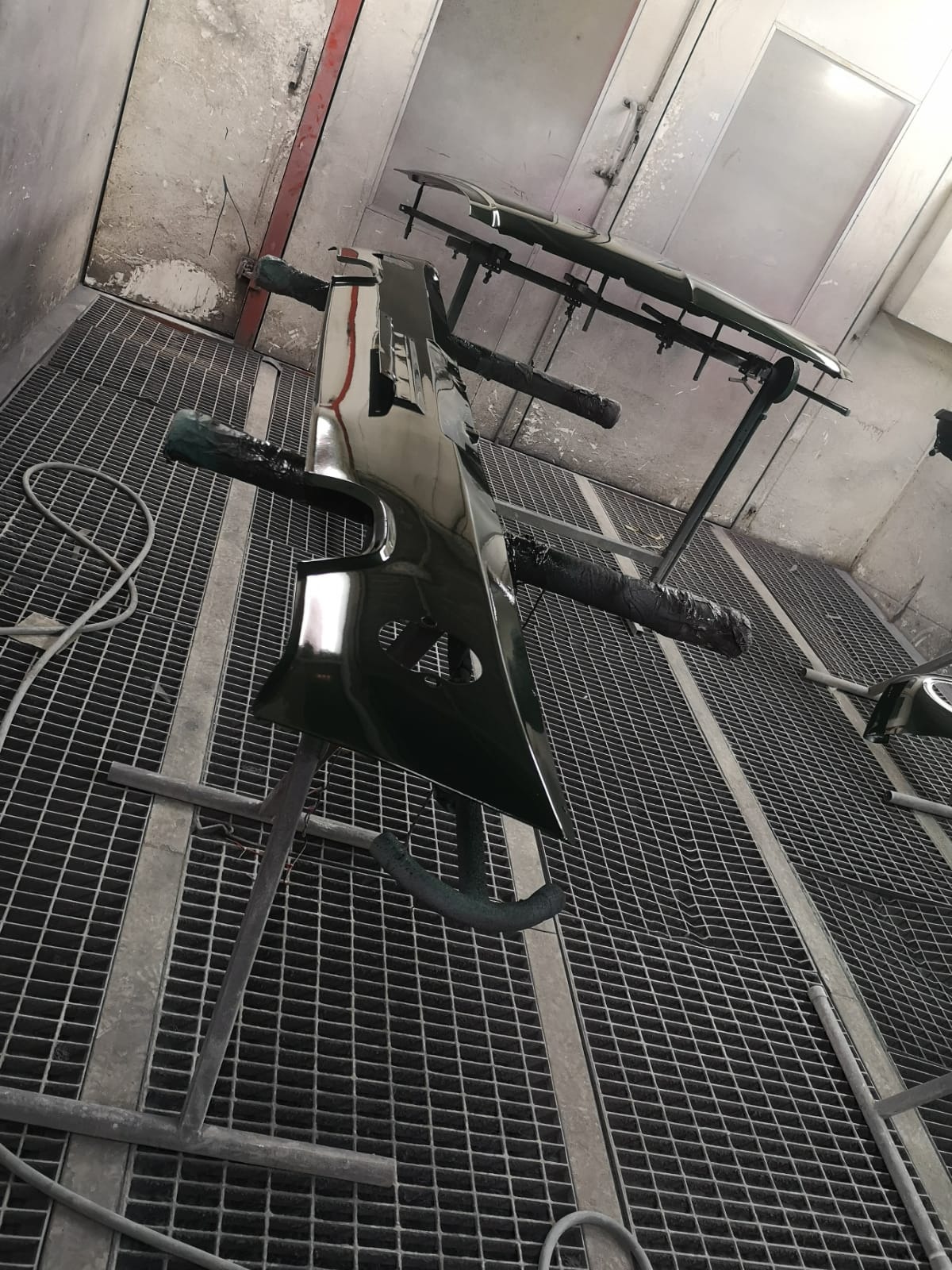 Elementy karoserii samochodowej, błotnik i zderzak, po lakierowaniu w kabinie lakierniczej, zawieszone na stojakach, widoczna krata podłogowa i ściany kabiny.