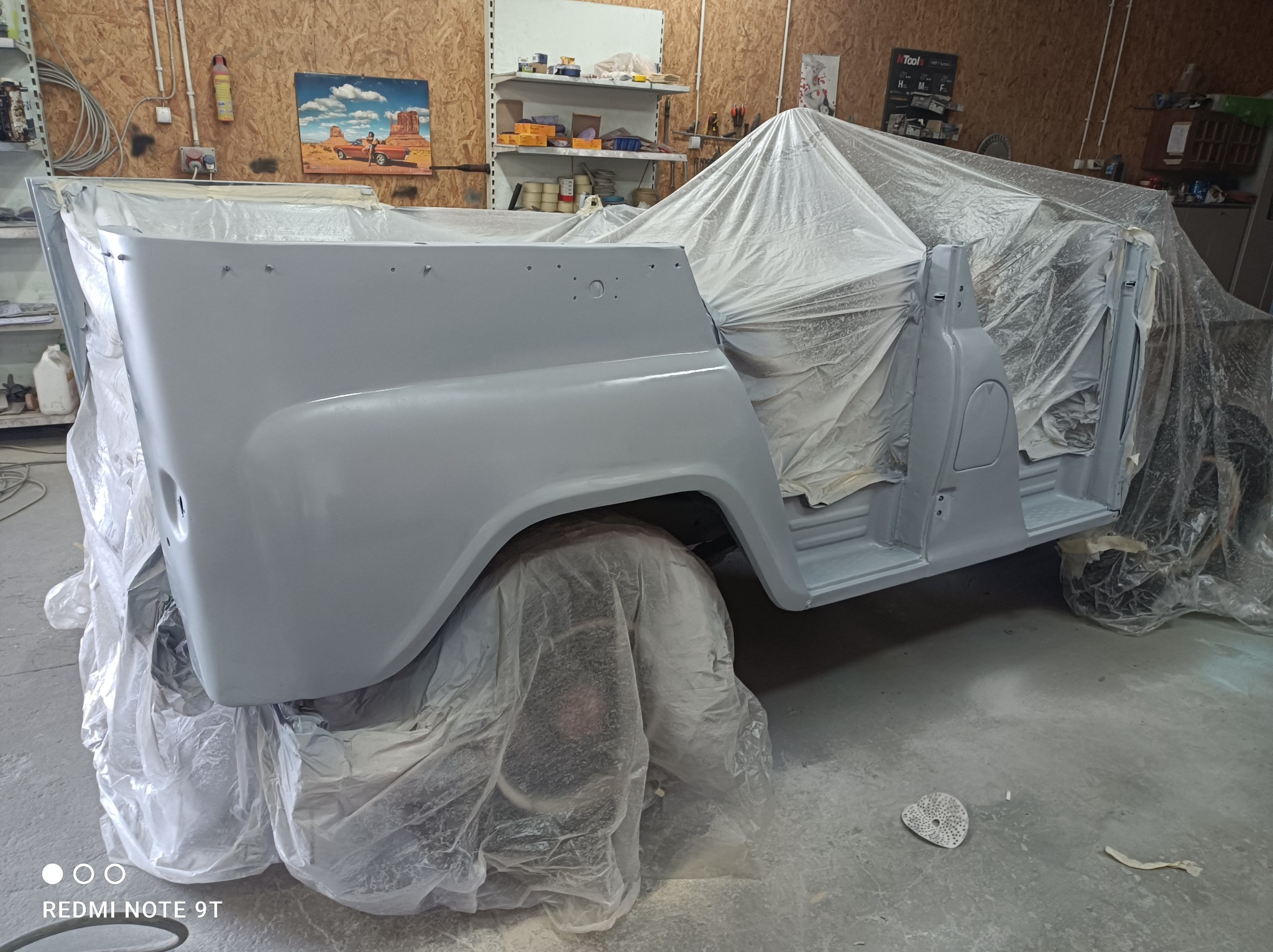 Szary, częściowo okryty folią samochód w trakcie lakierowania w warsztacie, widoczne drzwi i nadkola, na podłodze ślady po szlifowaniu.