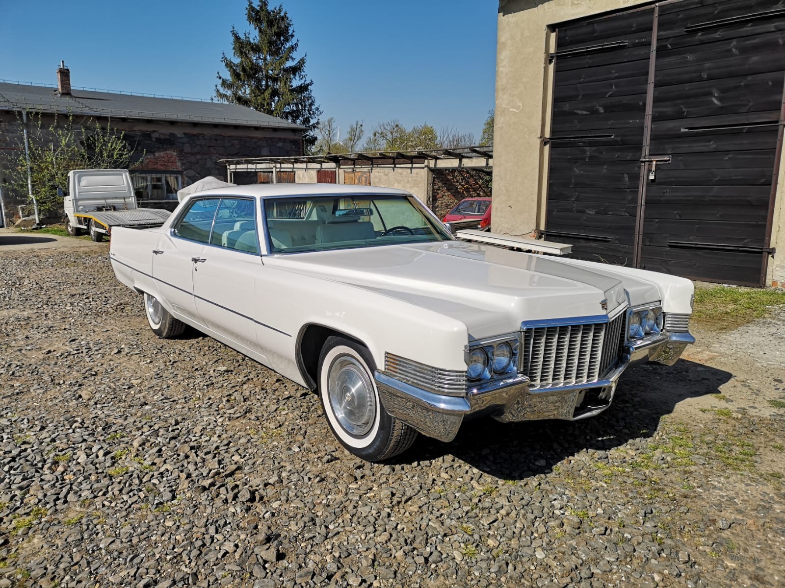 Biały, klasyczny Cadillac na żwirowym podłożu przed budynkiem z czarnymi, drewnianymi wrotami garażowymi.