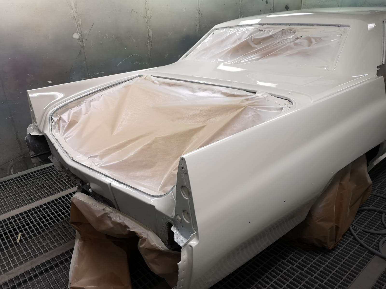 Klasyczny samochód Cadillac w trakcie lakierowania, zabezpieczony folią i papierem maskującym, widoczny fragment karoserii w jasnym odcieniu w kabinie lakierniczej.