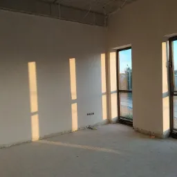 Wnętrze w trakcie wykańczania: ściany po gładziowaniu, widoczne ślady po pracach, surowy betonowy sufit i promienie słoneczne wpadające przez okna.