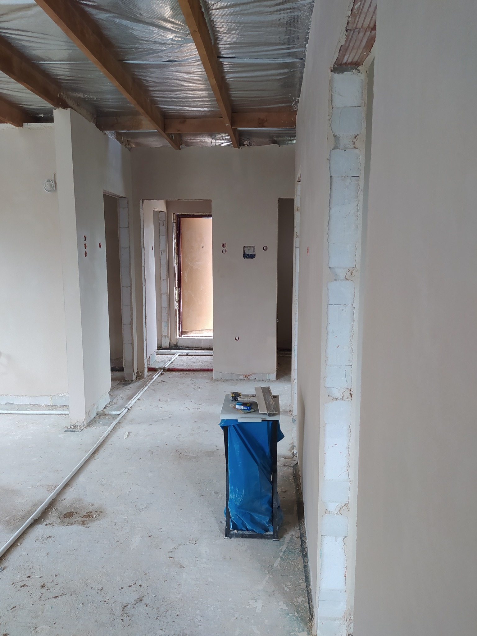 Wnętrze w trakcie remontu: widoczne ściany po nałożeniu gładzi, sufit z izolacją, prowizoryczny stolik z narzędziami w centralnej części kadru, instalacja elektryczna na podłodze.