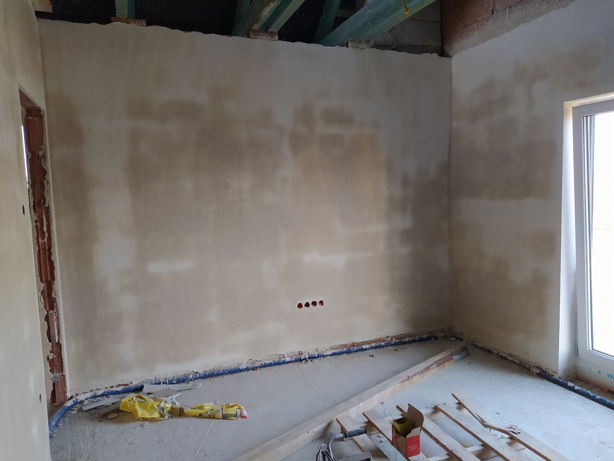 Wnętrze w trakcie prac wykończeniowych: ściany pokryte surowym tynkiem, widoczne otwory na gniazdka elektryczne, niebieska rurka przy podłodze i drewniane elementy konstrukcyjne na podłodze.