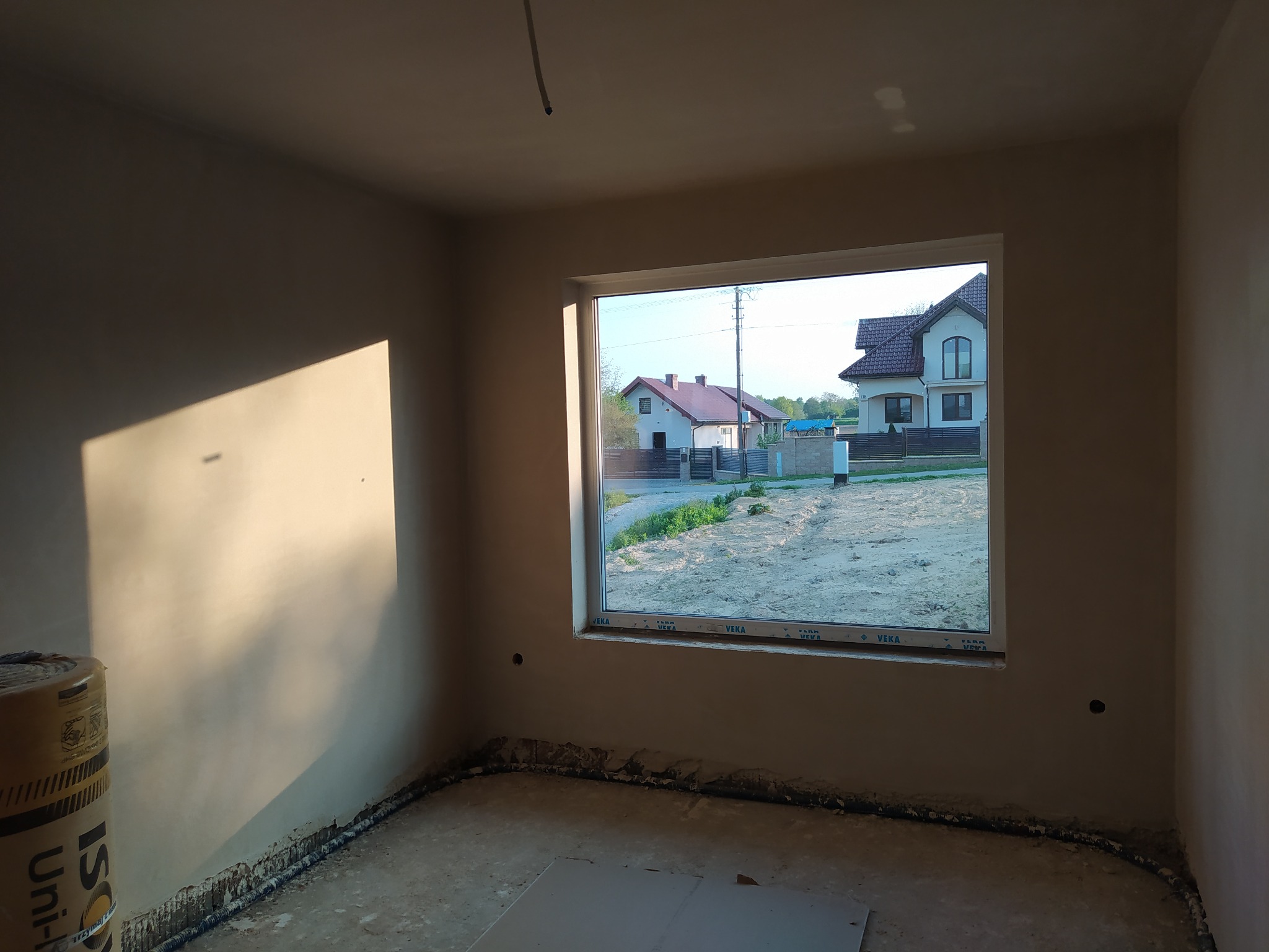 Wnętrze w trakcie prac wykończeniowych: ściany po nałożeniu tynku, widoczne okno z widokiem na domy jednorodzinne, rolka materiału izolacyjnego w rogu pomieszczenia, ślady prac na podłodze.