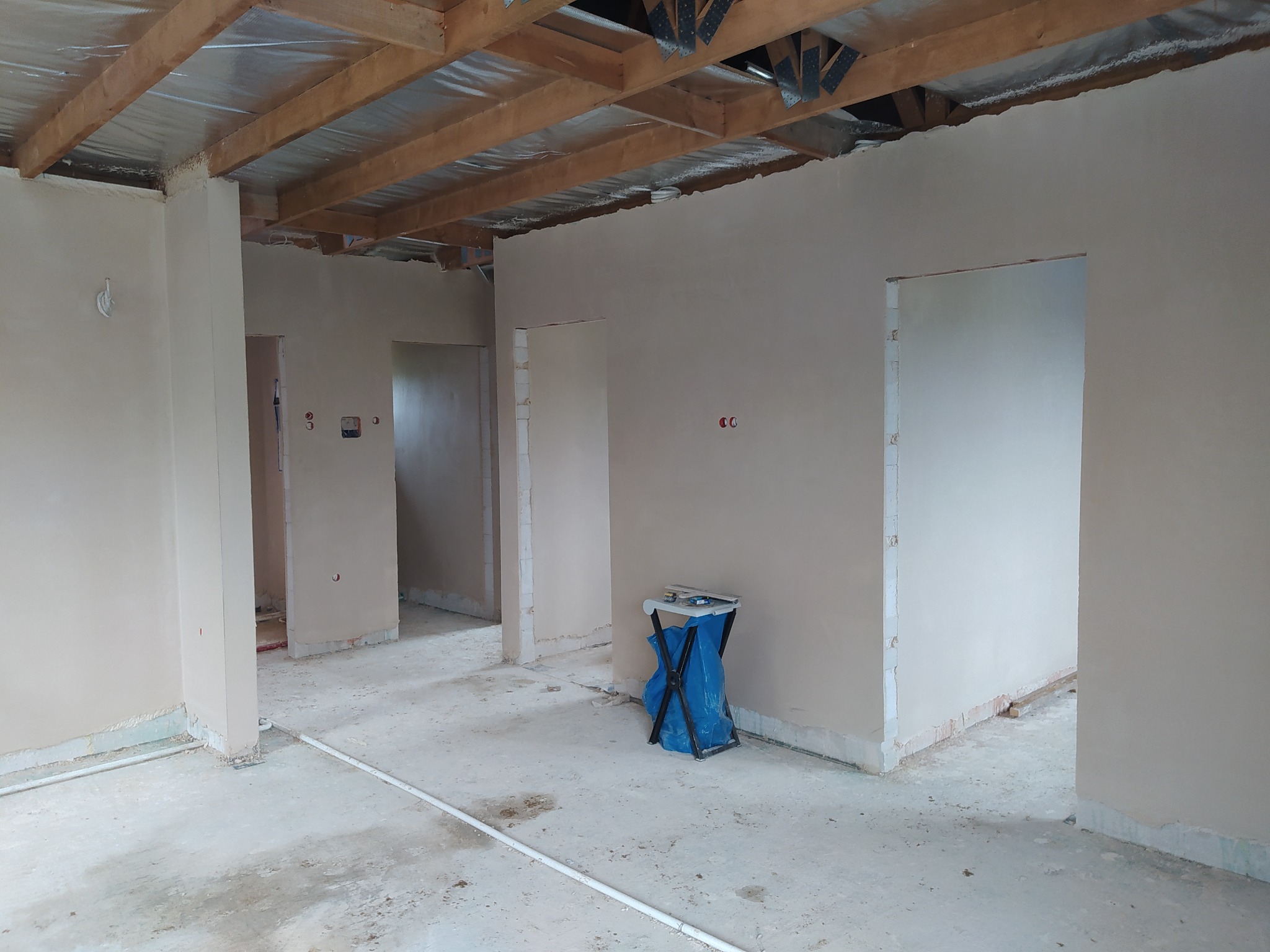 Wnętrze w stanie deweloperskim po nałożeniu tynków, widoczne surowe drewniane belki stropowe z folią izolacyjną, ściany pokryte świeżym tynkiem, na podłodze resztki materiałów budowlanych, w tle...