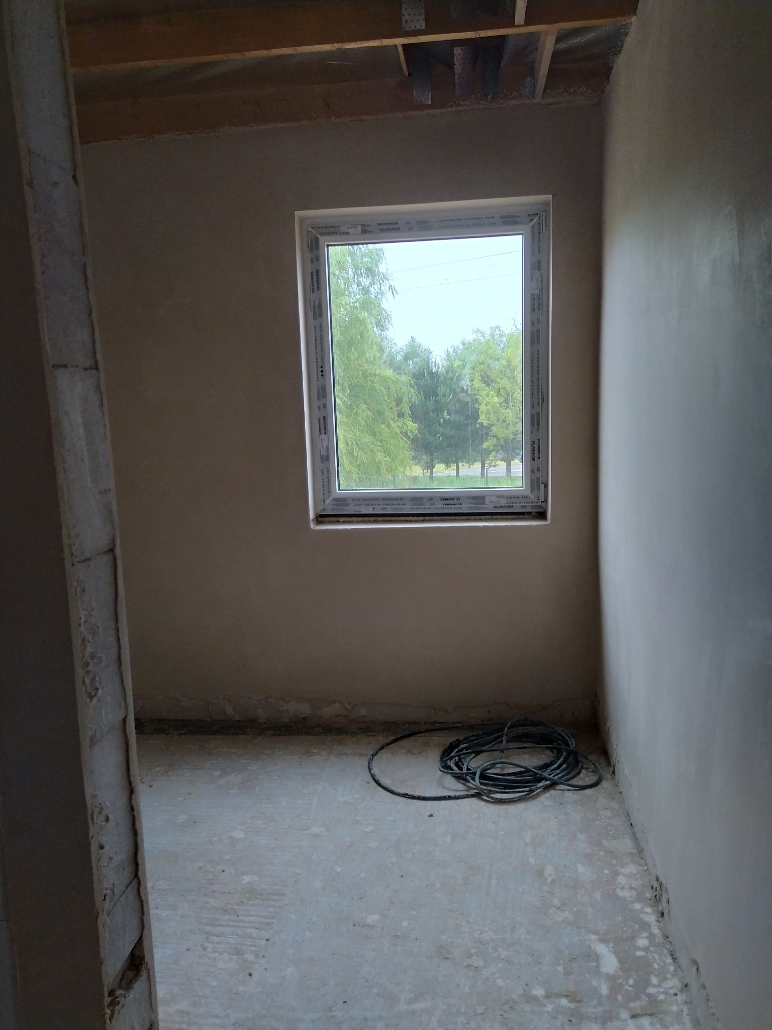 Surowe wnętrze pokoju po nałożeniu tynku, widoczne okno z widokiem na zieleń, zwój kabli na podłodze i surowe drewniane belki stropowe.