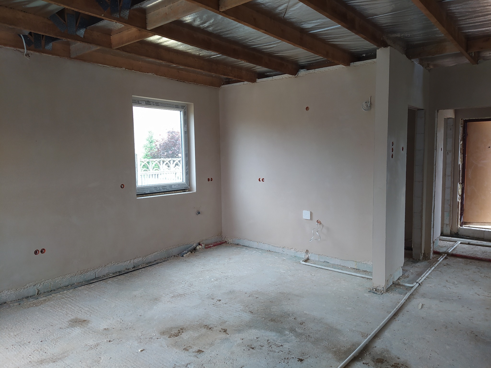 Wnętrze w trakcie remontu: ściany po nałożeniu gładzi, widoczne instalacje elektryczne i hydrauliczne, surowy sufit z izolacją, okno z widokiem na zieleń.