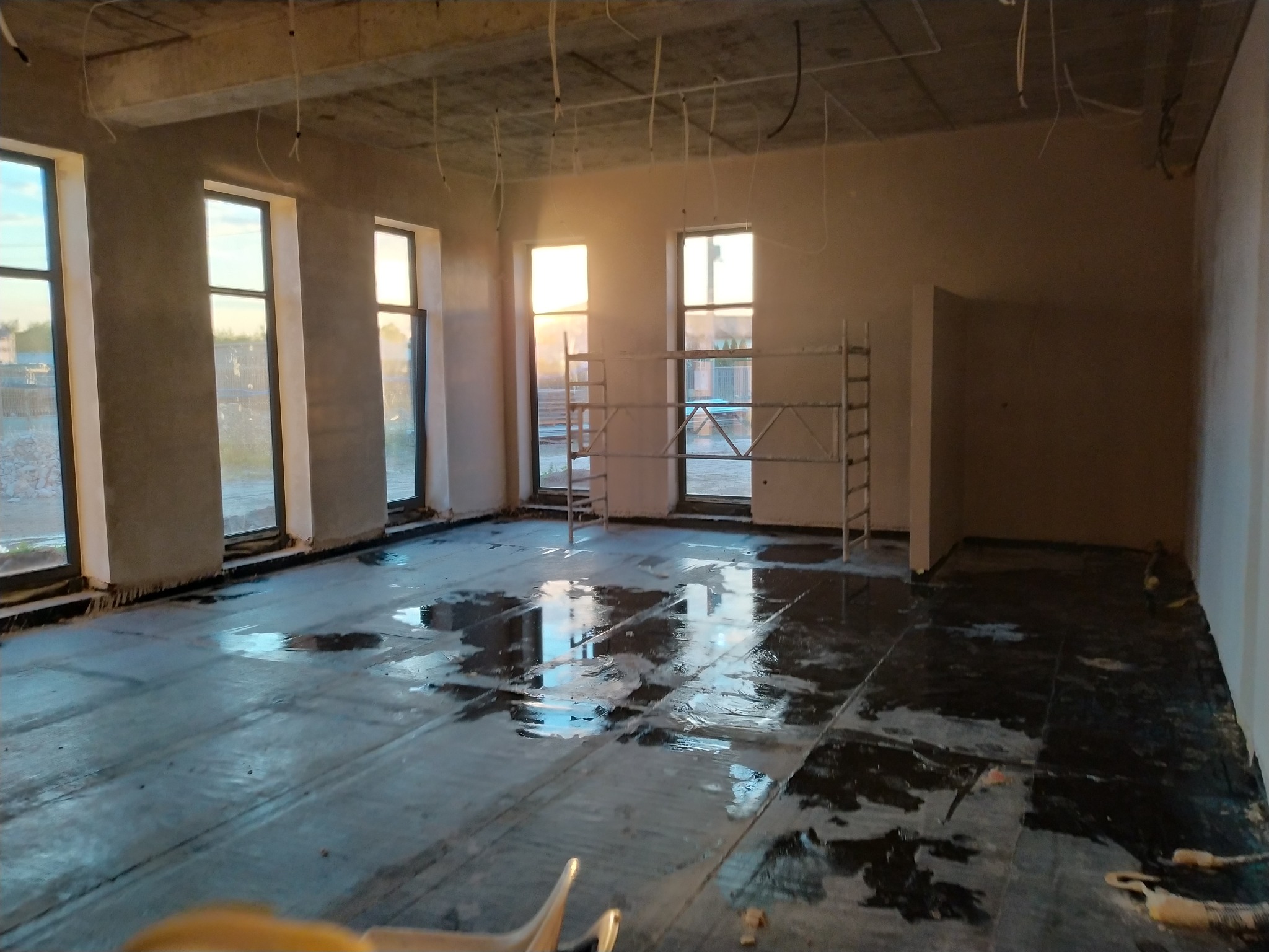 Wnętrze w stanie surowym z trzema oknami i rozstawionymi rusztowaniami, podłoga zabezpieczona folią, widoczne kable zwisające z sufitu.