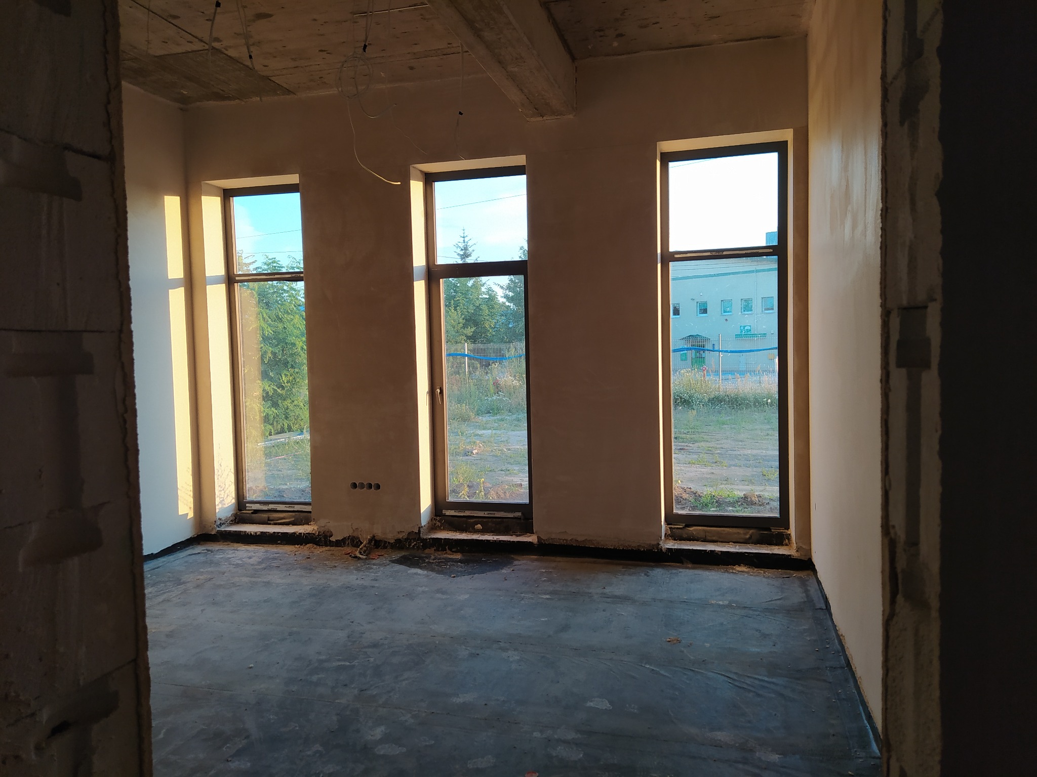 Surowe wnętrze z trzema oknami, ściany po nałożeniu tynku, widoczne przewody elektryczne w suficie i folia ochronna na podłodze.