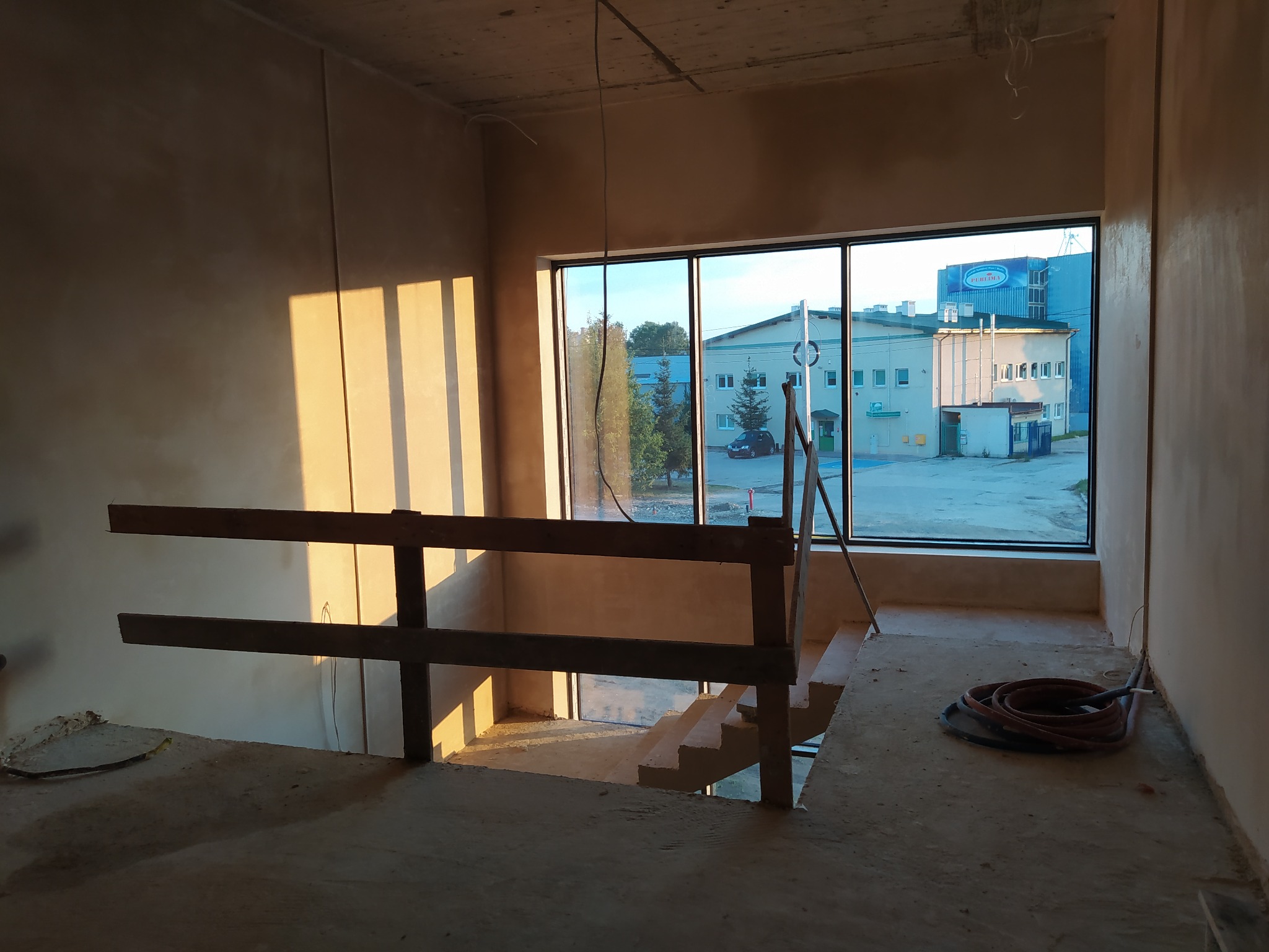 Surowe, oświetlone promieniami słońca wnętrze w trakcie prac wykończeniowych; ściany pokryte świeżym tynkiem, widoczne schody, prowizoryczna barierka i zwój węży na podłodze, okno z widokiem...