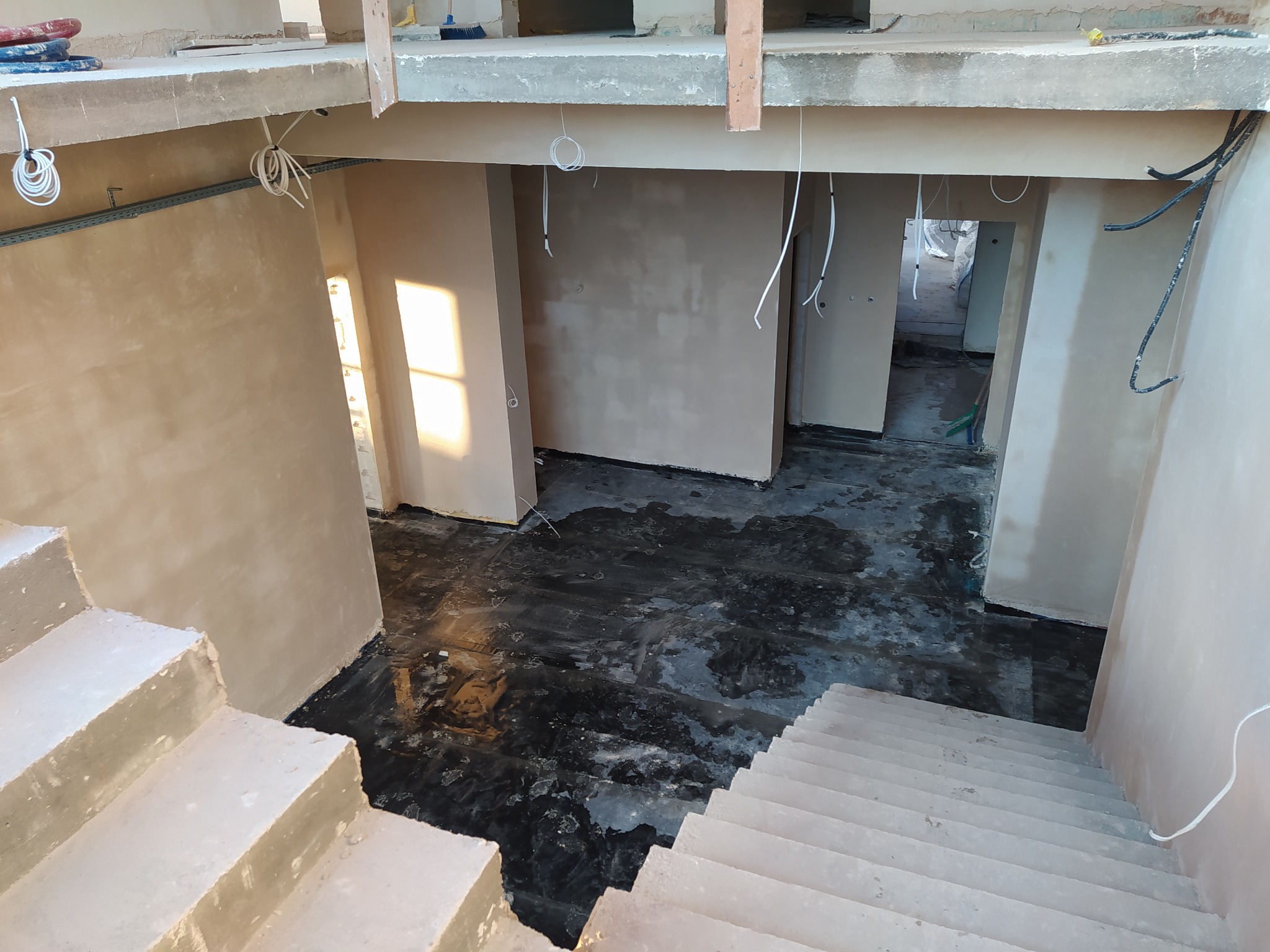 Wnętrze domu w trakcie remontu: ściany po tynkowaniu maszynowym, zabezpieczona podłoga, schody w surowym stanie, wiszące przewody elektryczne.