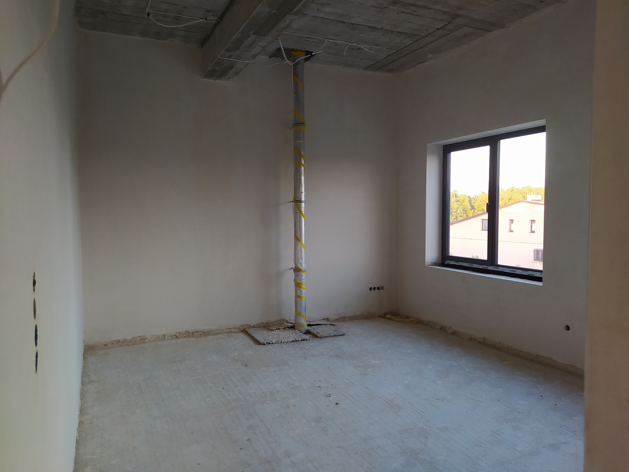 Surowe wnętrze pokoju w stanie deweloperskim z widocznymi śladami prac wykończeniowych na ścianach i podłodze, zabezpieczona rura pionowa na środku pomieszczenia, widok z okna na budynek i zieleń.