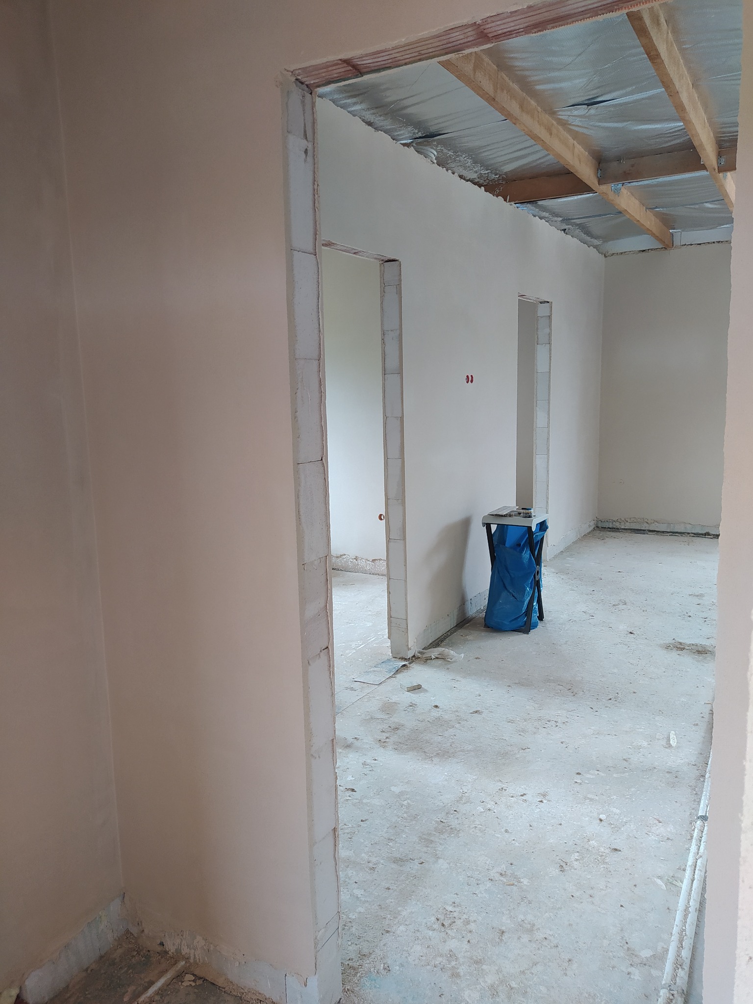 Wnętrze w trakcie remontu: ściany pokryte świeżo nałożoną gładzią, widoczne fragmenty ocieplenia pod stropem, surowy beton na podłodze i niebieski worek na śmieci, ujęcie przez futrynę drzwiową.