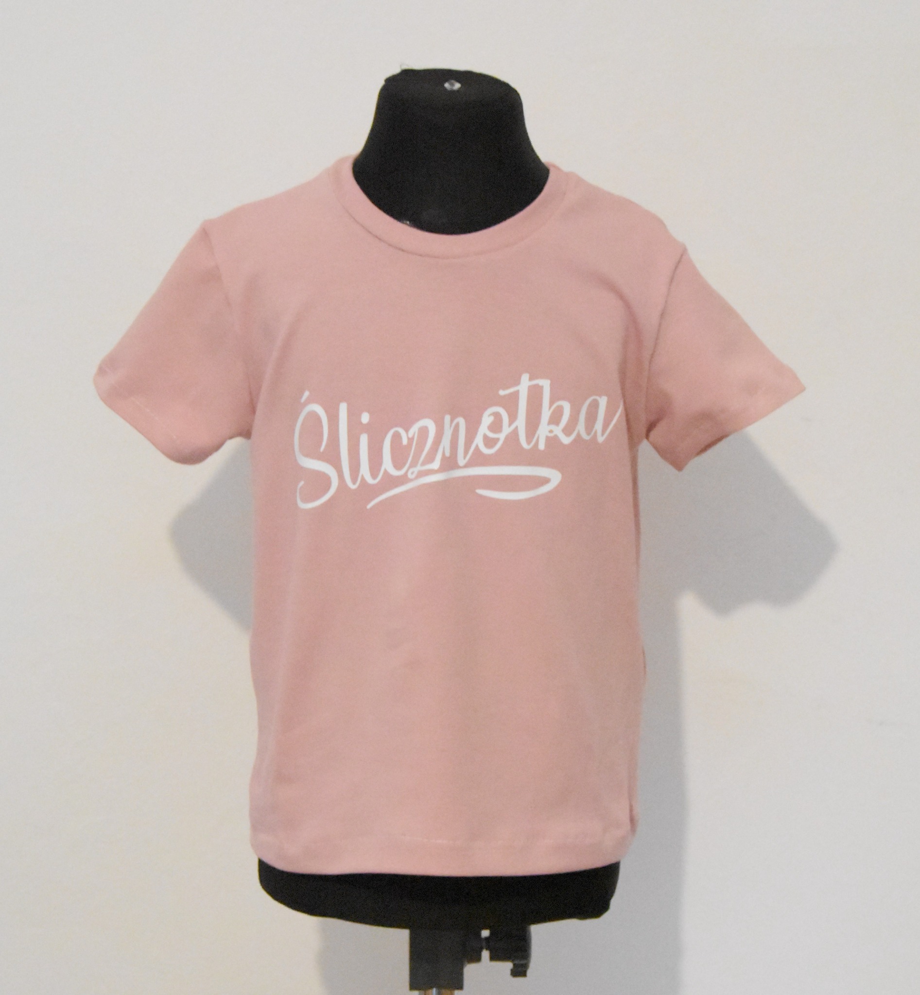 Różowa koszulka dziecięca z napisem 'Ślicznotka' na czarnym manekinie, widok z przodu na jasnym tle.