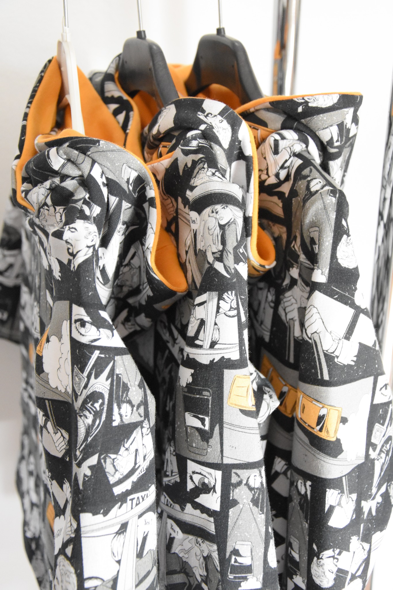 Trzy czarno-białe bluzy z kapturem, wykończone pomarańczową podszewką, wiszące na czarnych wieszakach. Bluzy zdobione komiksowym wzorem z postaciami i dymkami.