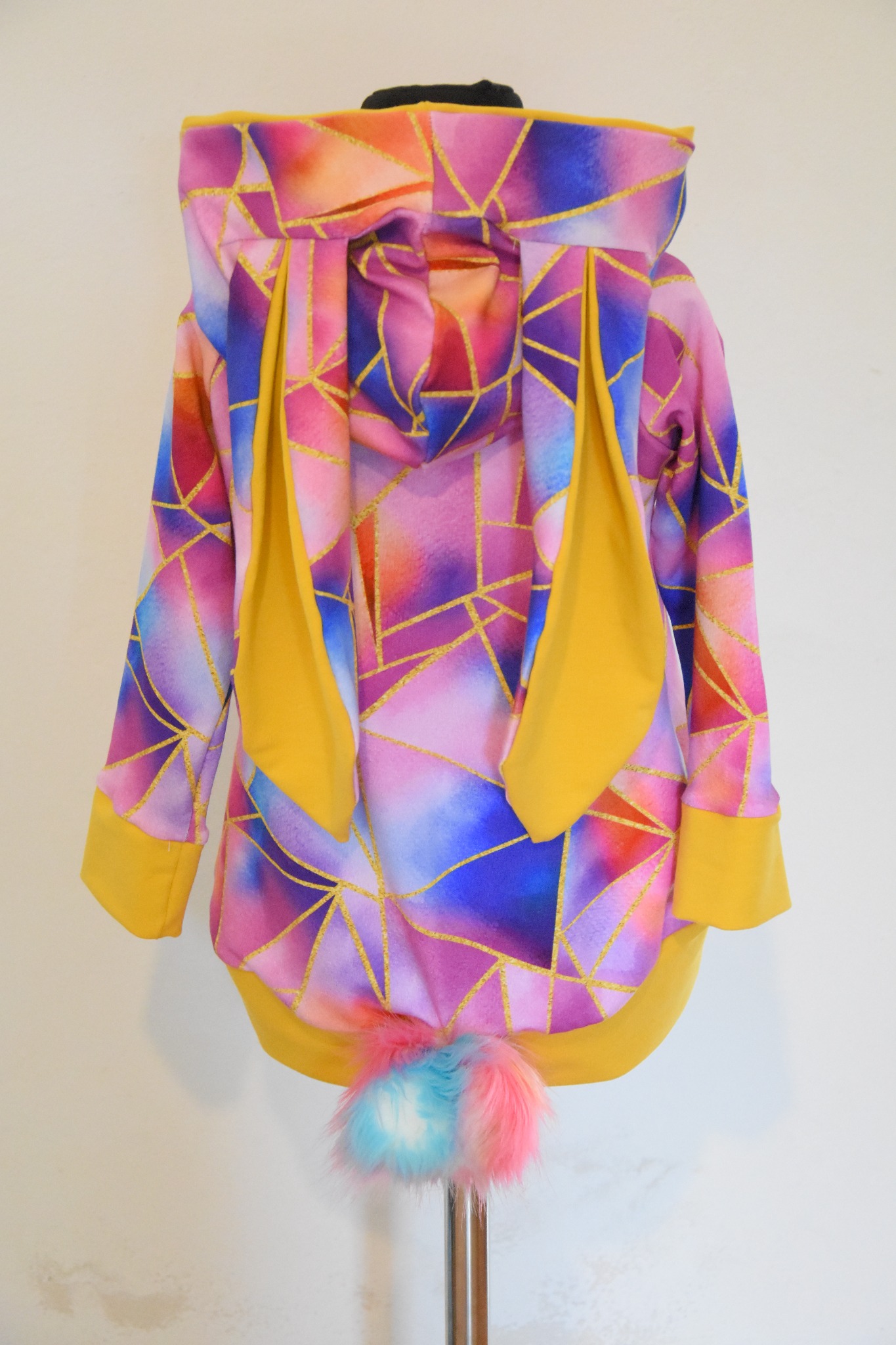 Kolorowa bluza z kapturem, uszami królika i pomponem ogonem, wisząca na wieszaku, wzór geometryczny z brokatowymi liniami.