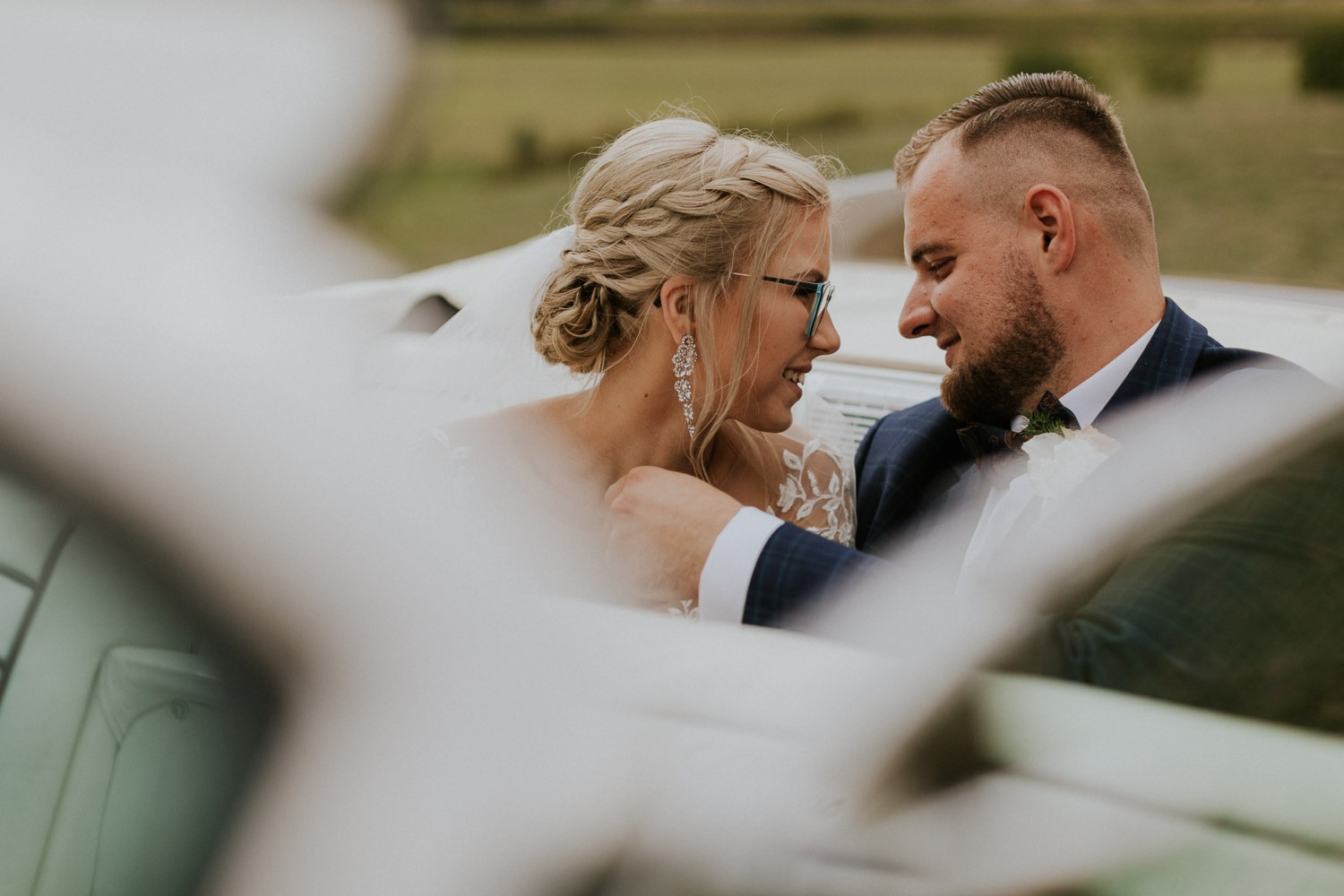 Uśmiechnięta para młoda w kabriolecie: panna młoda w okularach z upiętym blond warkoczem i długimi kolczykami, pan młody w granatowym garniturze w kratę i muszce, zieleń w tle.