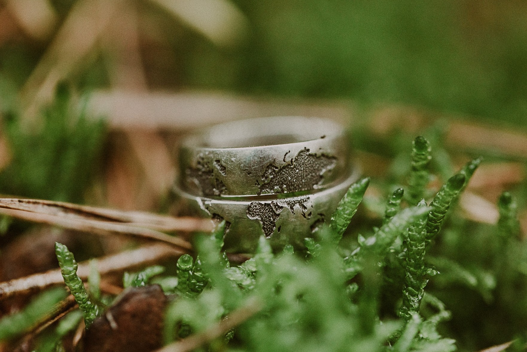 Dwa srebrne obrączki ślubne z grawerowaną mapą świata, ułożone na mchu i igliwiu.