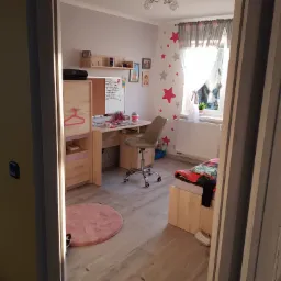 Widok na pokój dziecięcy z biurkiem, krzesłem na kółkach, łóżkiem i dywanem, przez otwarte drzwi z widocznym fragmentem innego pokoju.