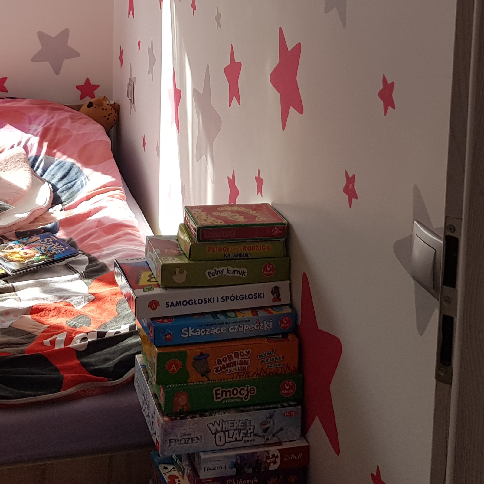Pokój dziecięcy z łóżkiem w różowe wzory i stertą gier planszowych ułożonych obok ściany ozdobionej naklejkami w kształcie gwiazd.