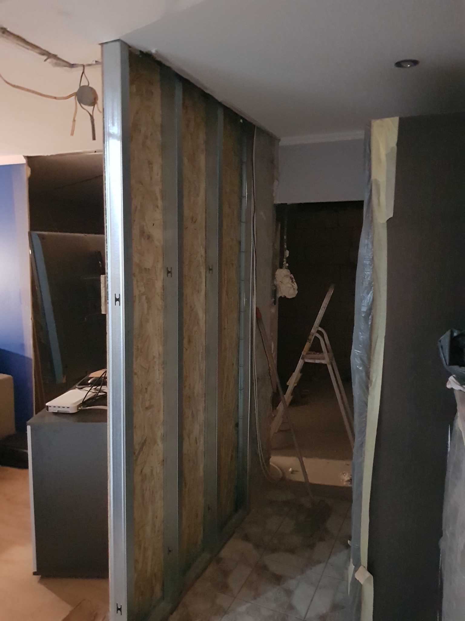 Wnętrze w trakcie remontu: konstrukcja ścianki działowej z metalowych profili wypełnionych płytą OSB, widoczne instalacje elektryczne, drabina w tle, przygotowania do malowania.