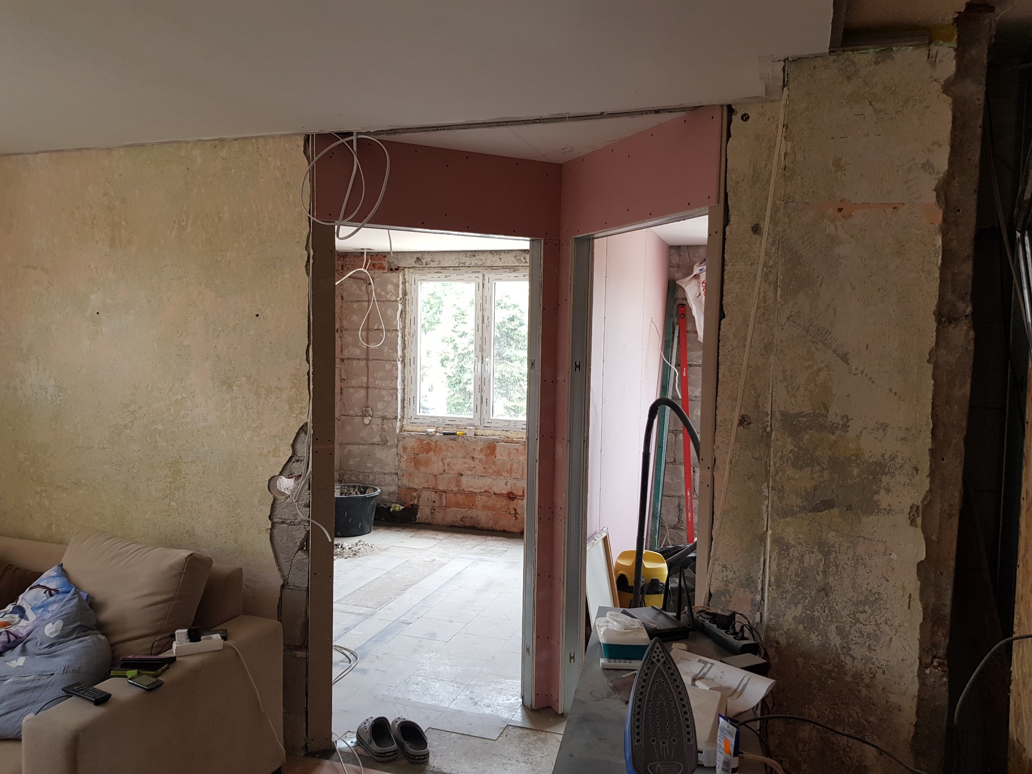 Wnętrze mieszkania w trakcie demontażu ścian działowych, widoczne odsłonięte cegły, przewody elektryczne, fragment sofy i sprzęty budowlane.