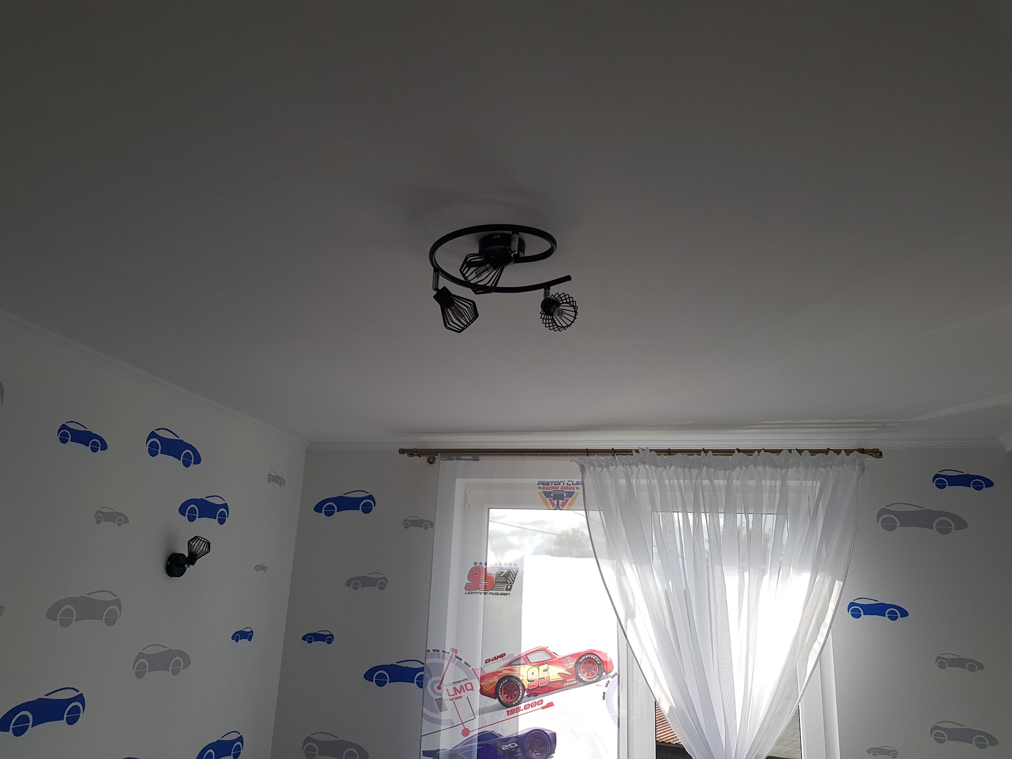 Pokój dziecięcy z motywem samochodów: białe ściany z naklejkami aut w odcieniach szarości i niebieskiego, okno z białymi firankami z nadrukiem samochodów wyścigowych, czarna lampa sufitowa z trzema...