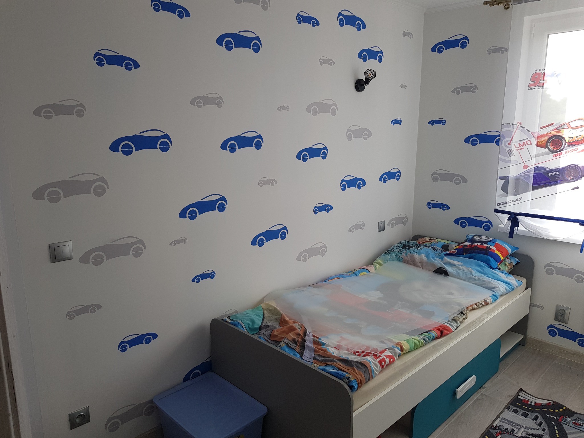Pokój dziecięcy z łóżkiem, ścianą ozdobioną naklejkami samochodów w kolorach niebieskim i szarym, zasłoną z motywem wyścigowym oraz niebieskim pojemnikiem na zabawki.
