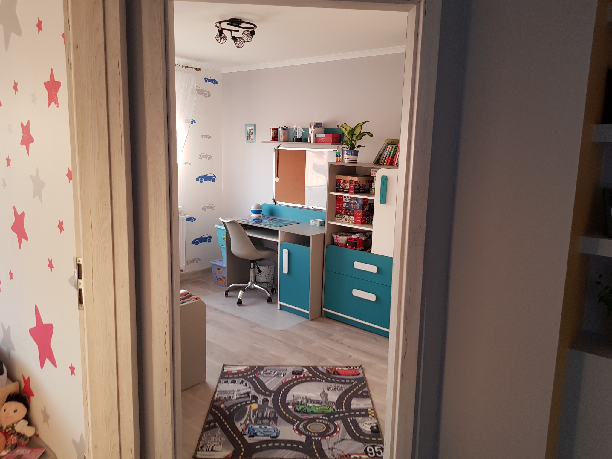Pokój dziecięcy z biurkiem, regałem i dywanem z motywem toru wyścigowego, widoczny przez otwarte drzwi z dekoracją w gwiazdki.