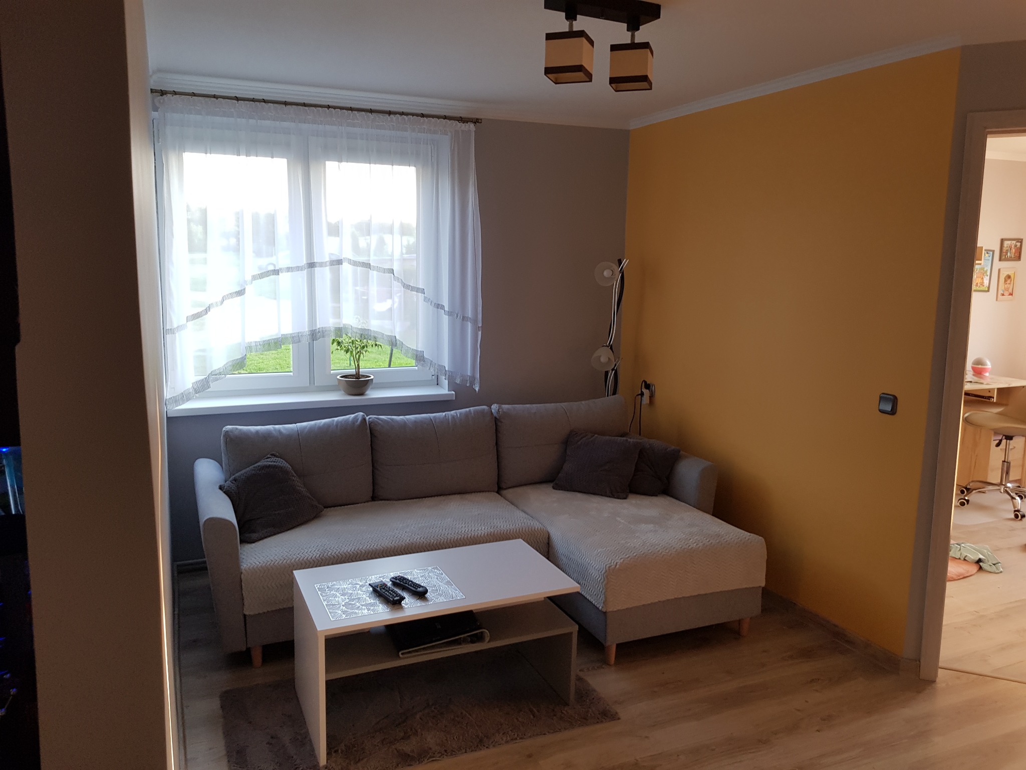 Jasny salon z szarą narożną sofą, białym stolikiem kawowym z pilotami, żółtą ścianą i oknem z firanką, widok na wnętrze przez otwarte drzwi.