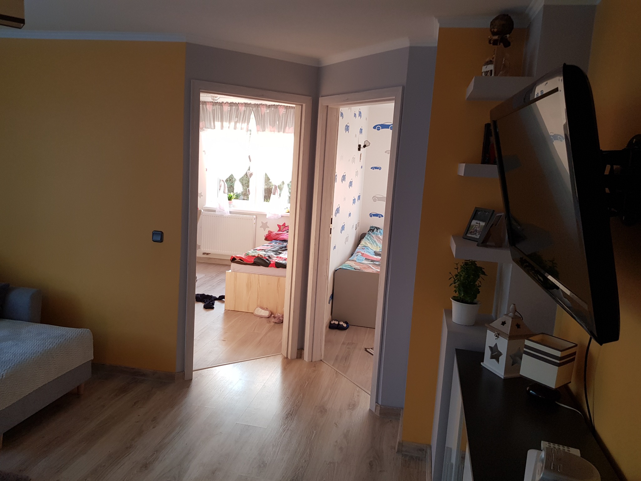 Salon z widokiem na dwa pokoje dziecięce przez otwarte drzwi, z panelami podłogowymi i żółtymi ścianami, półki z dekoracjami i telewizorem.