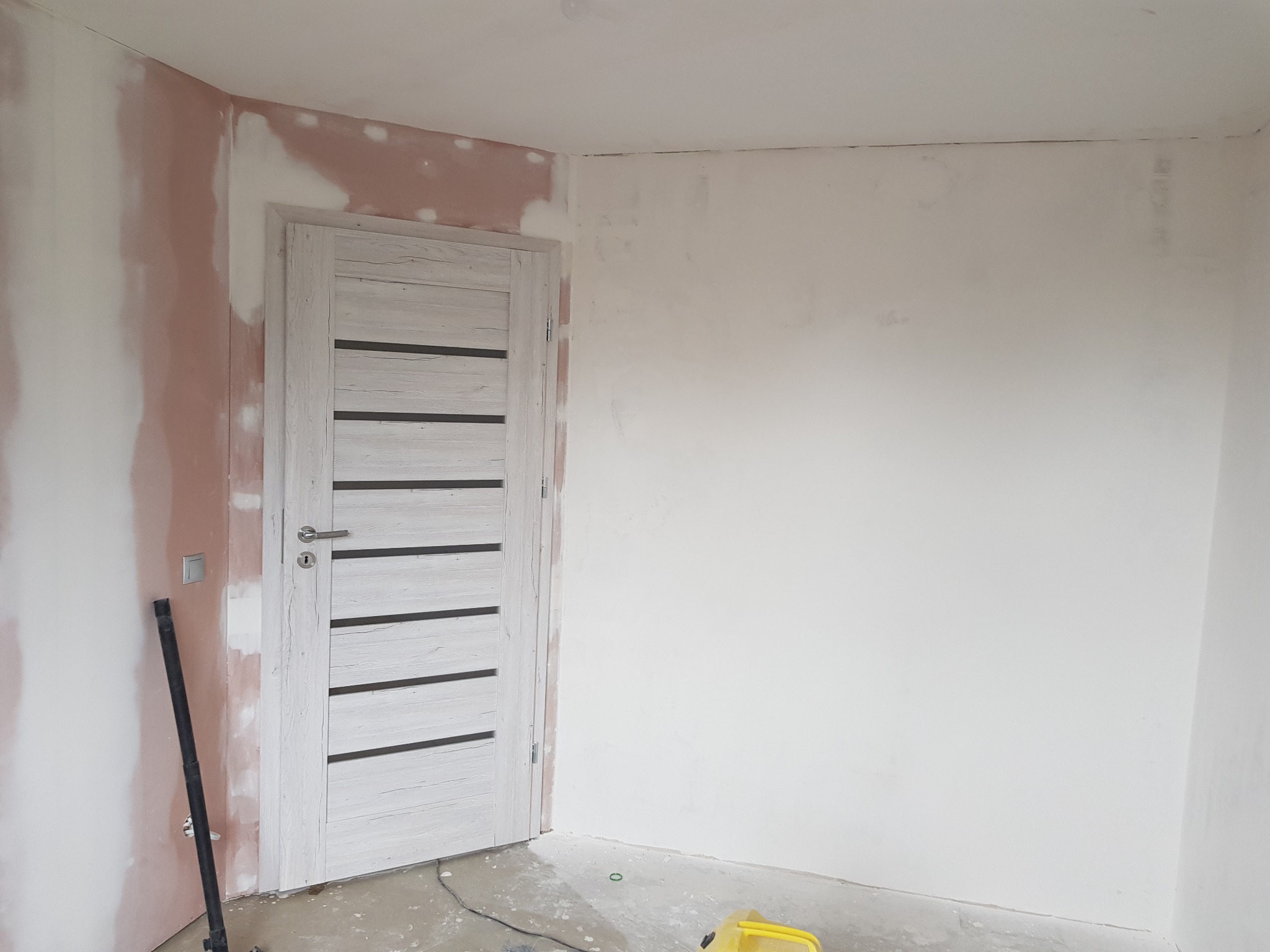 Pokój w trakcie prac wykończeniowych: ściany częściowo zagruntowane, widoczna szpachla, zamontowane białe drzwi wewnętrzne z poziomymi, ciemnymi wstawkami, na podłodze widoczne ślady prac, narzędzia.