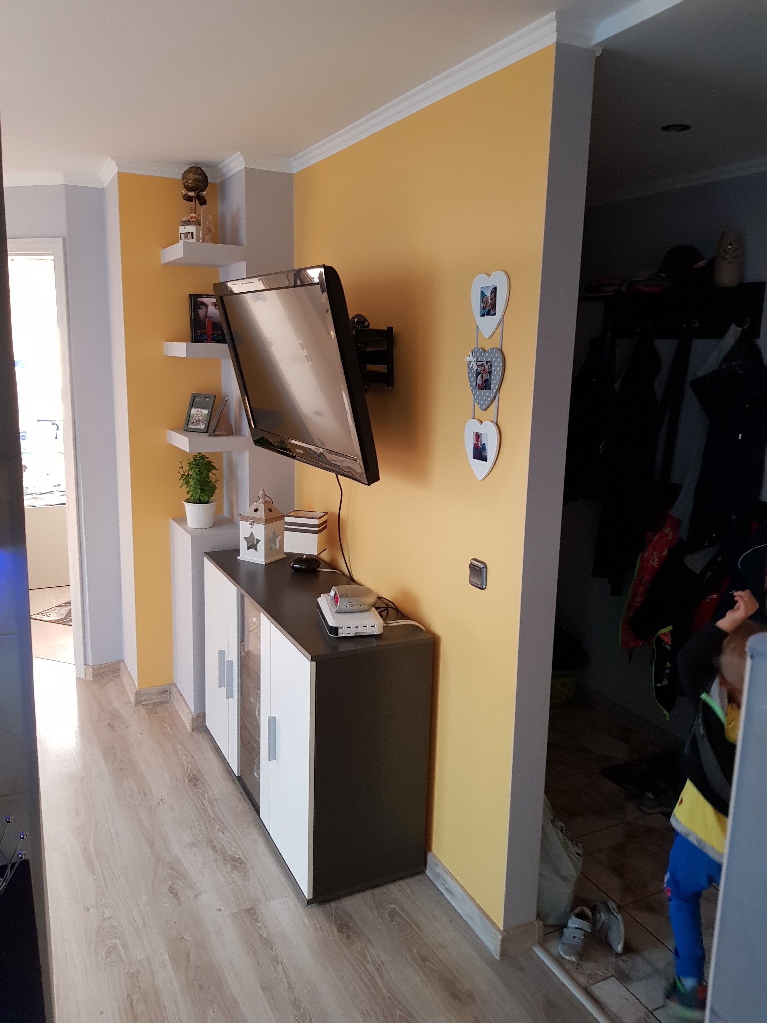 Salon po odświeżeniu: żółta ściana, podwieszony telewizor, biało-czarna komoda, drewnopodobne panele podłogowe, dekoracje na ścianie w kształcie serc i półki z ozdobami.